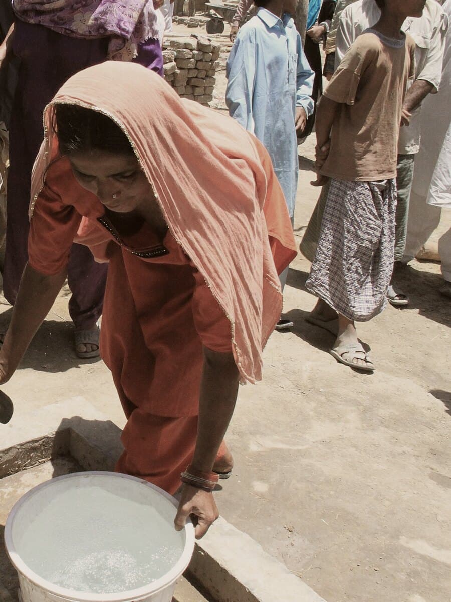 A woman drawing water from a village hand pump as children wait